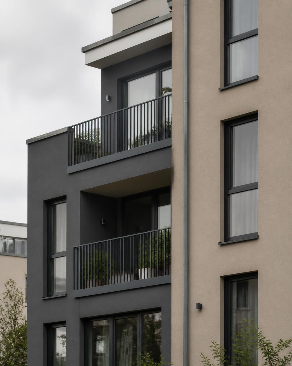 Fassade eines modernen Mehrfamilienhauses mit anthrazitfarbenem und beigem Putz, dunklen Fensterrahmen und bepflanzten Balkonen mit Metallgeländer